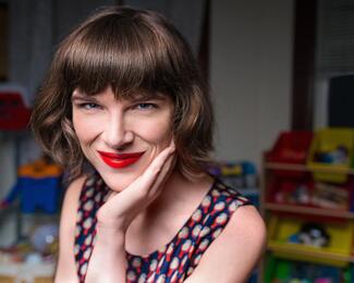 Photo of light skinned woman with dark red hair and bright red lips looking at camera. Smiling with hand under chin. Background looks like a room with kids toys