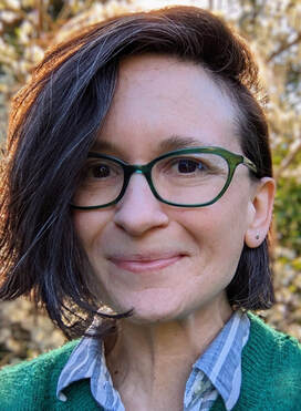 headshot of light skinned woman with dark hair to her chin. She is wearing glasses and a green sweater. Trees behind her